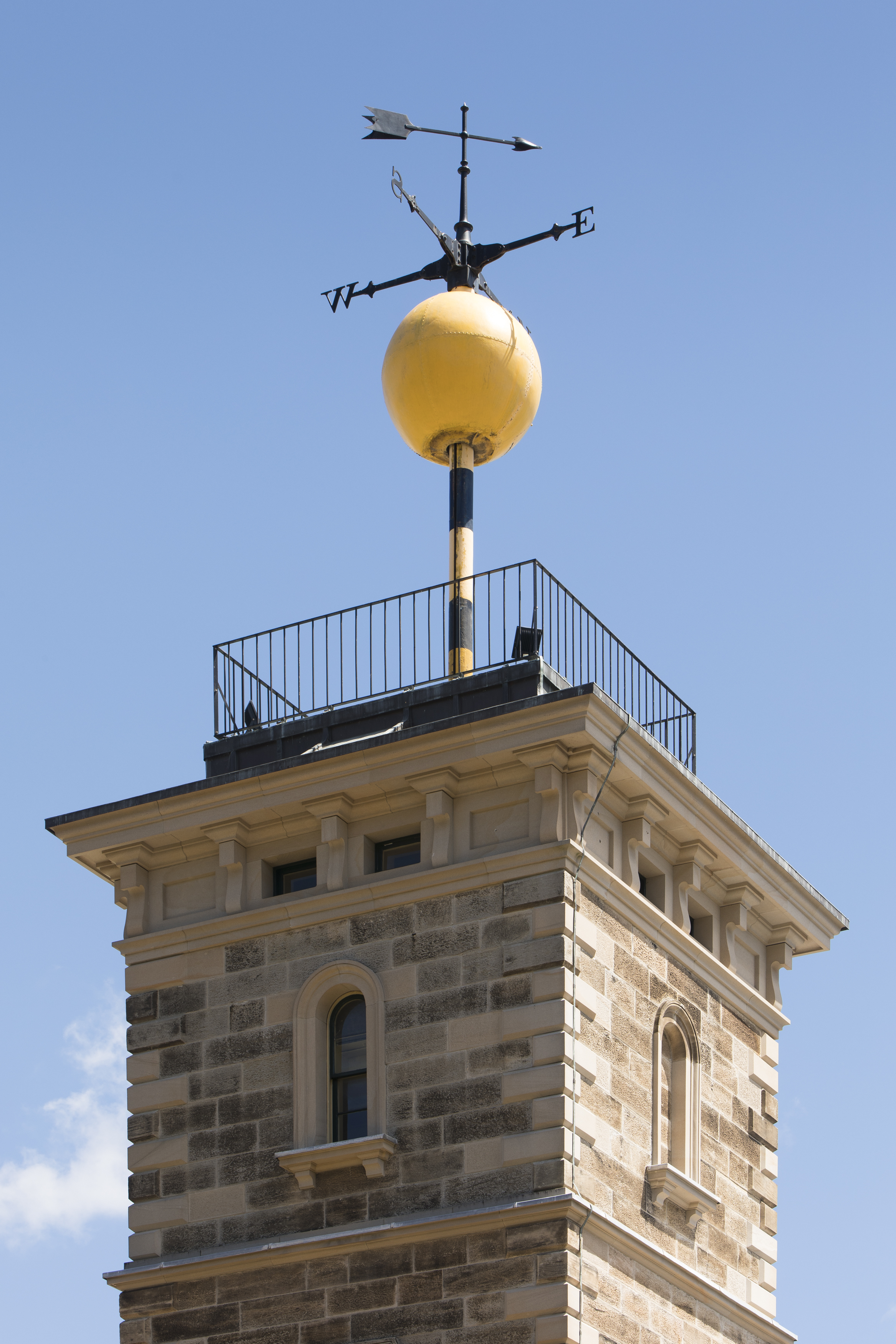 A large yellow metal call sitting on top of a sandstone tower with a weather vane above it.
